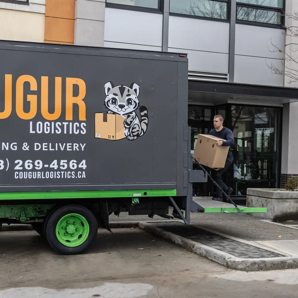 A delivery professional carries a cardboard box up the ramp of a Cougur Logistics truck parked outside a modern building.