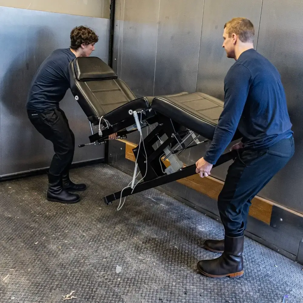 Two employees carefully maneuver a medical chair inside a spacious elevator with stainless steel walls.