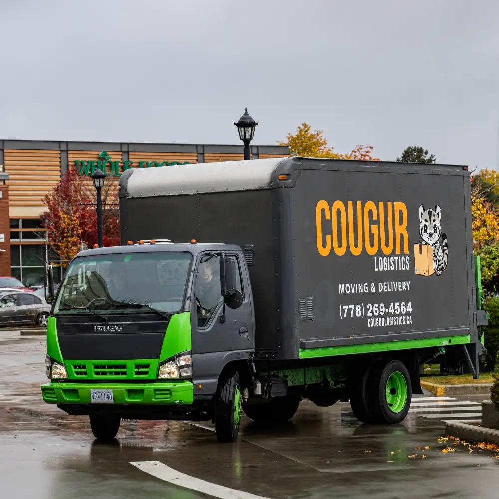 A green and black Isuzu delivery truck with "Cougur Logistics" branding parked near a Whole Foods Market.