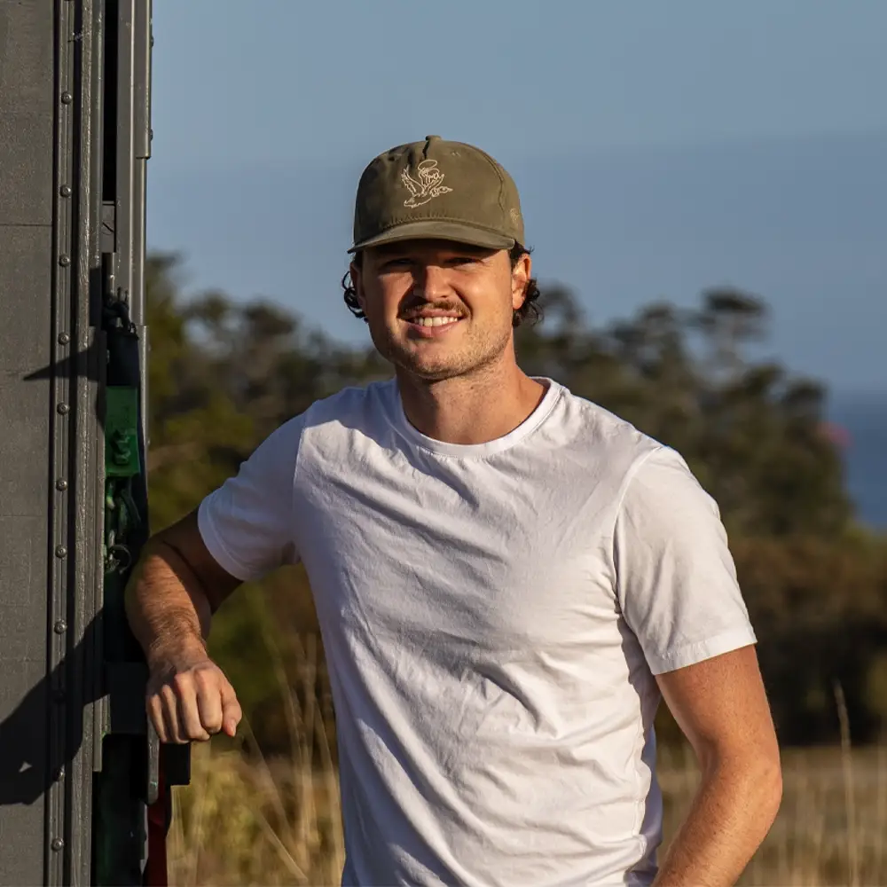 Headshot of Oliver Cougur, owner-operator of Cougur Logistics, leaning against his truck with the ocean and trees in the background.